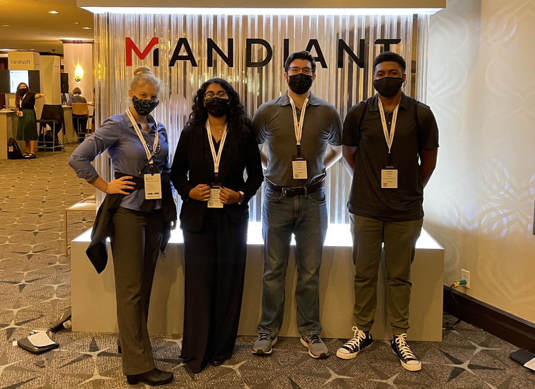 Four students standing in front of a vendor booth in a large conference room.