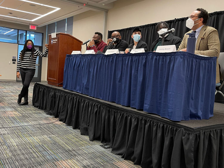 A group of men sit on a stage at a table with a blue table cloth with a woman standing beside the stage, moderating the discussion.