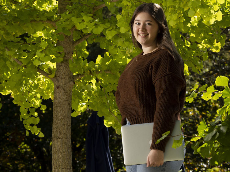 A female student stands outside holding a laptop.