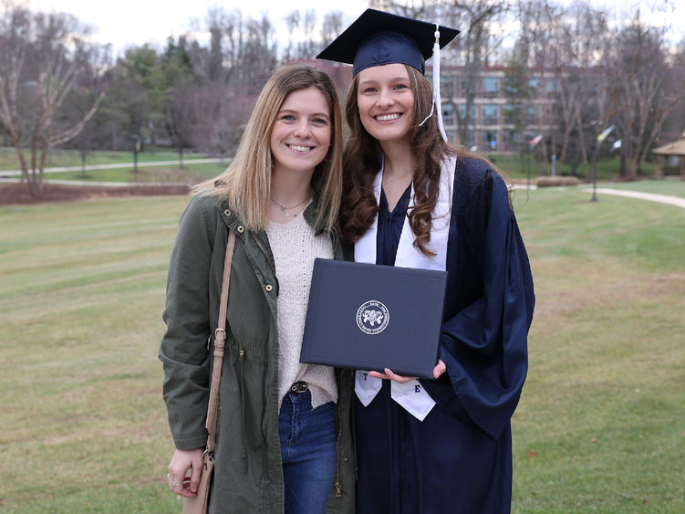 Two women stand outdoors. One is wearing a graduation cap and gown and holding a diploma cover.